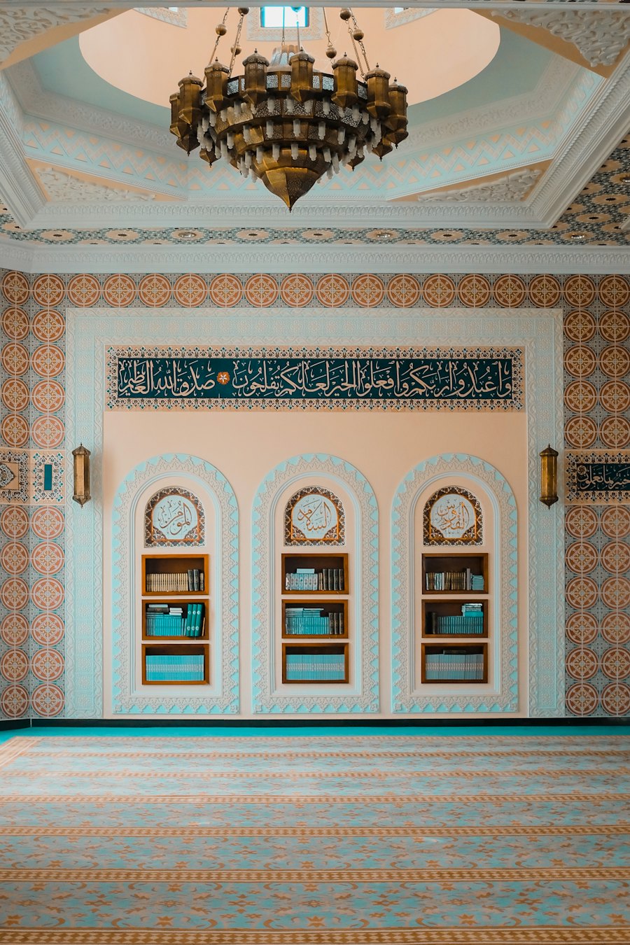A mosque interior with a brass chandelier, tiled walls and shelves of books under three arches